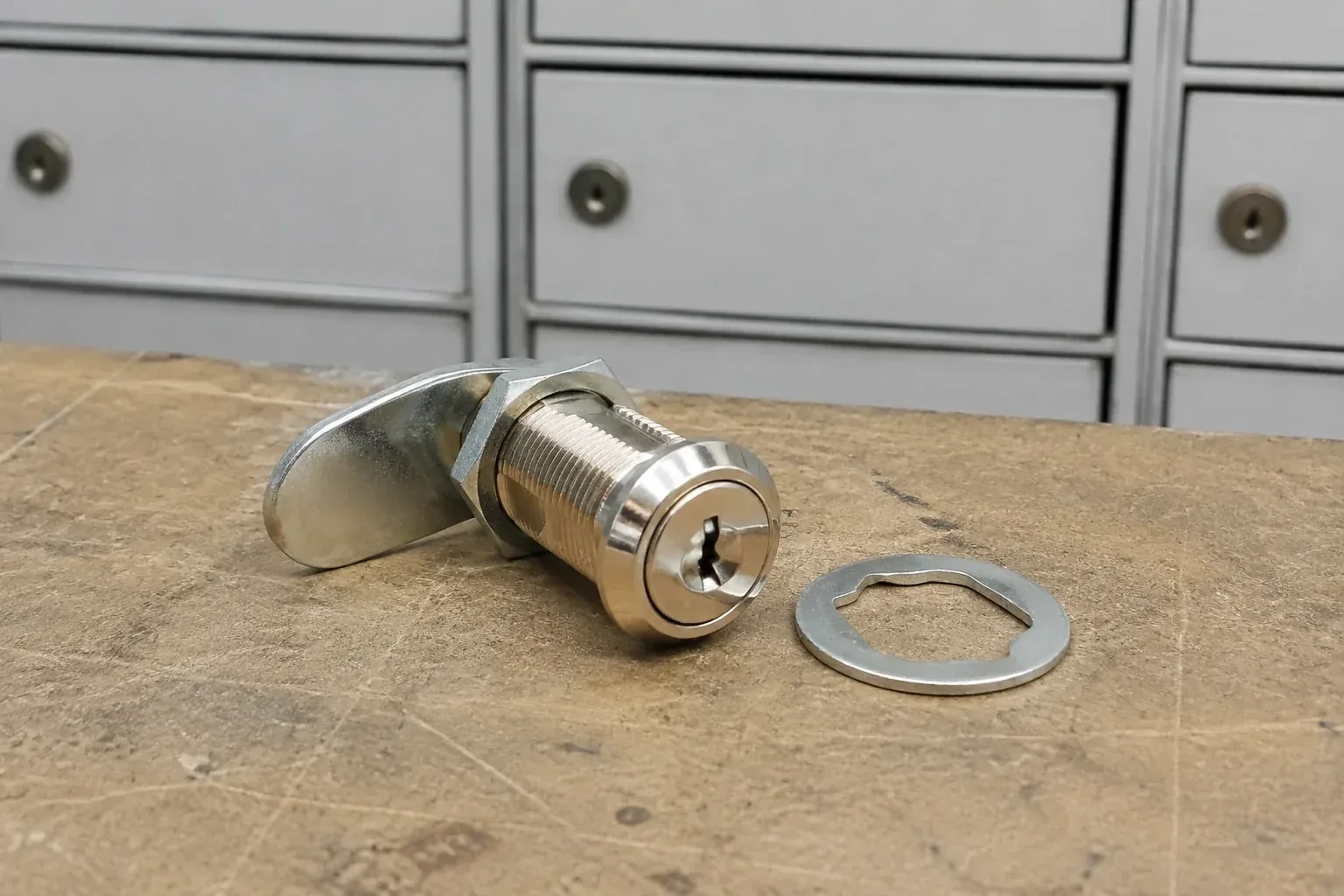 Detached threaded cam lock and mounting washer on a workbench in front of closed mail compartments. — Mailbox Lock Replacement, Hamilton