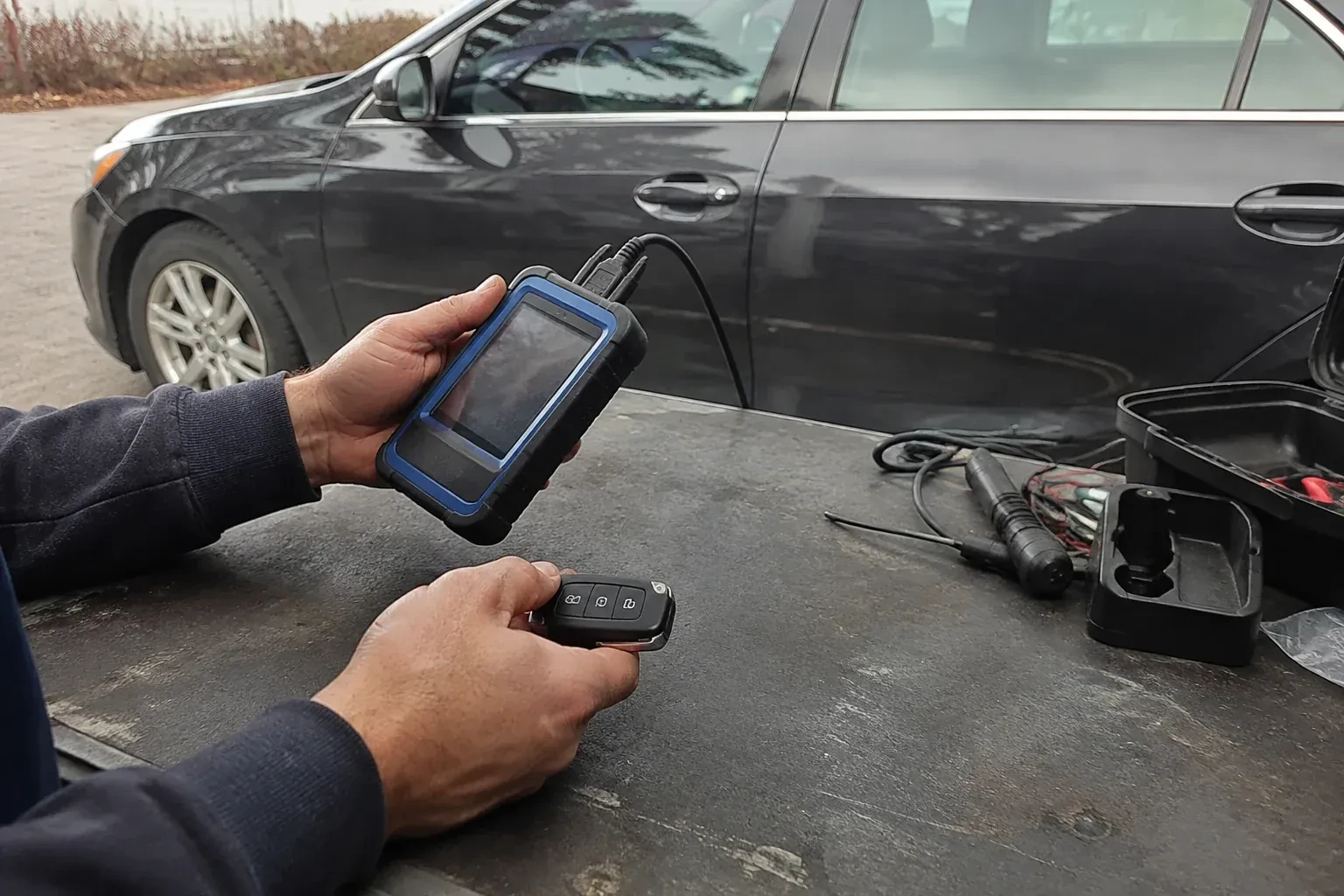 Hands holding a blue-edged electronic device and car-key fob at a worktable beside a parked car. — Transponder Key Programming, Cherry Heights