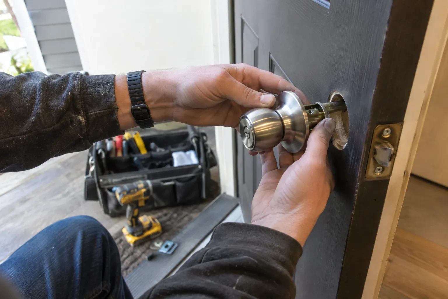 Hands fitting a steel door knob into the bore of a dark door, with a yellow drill and tool bag behind.